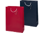 Duas sacolas de presente, uma vermelha e uma azul marinho, com alças de corda branca e etiquetas.