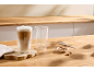 Dois copos, um com café latte, numa bancada de madeira com biscoitos e uma colher.