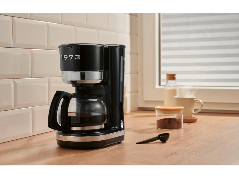 Máquina de café preta com jarra cheia de café, ao lado café moído, leite e uma caneca.
