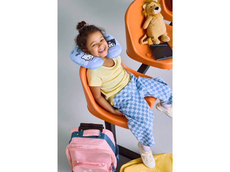 Menina feliz com almofada de viagem, mala e leão de peluche numa área de espera.