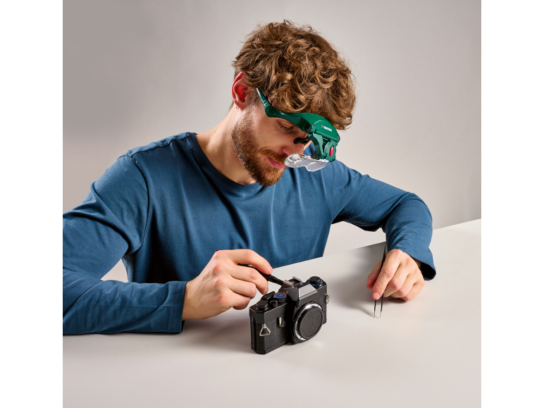 Homem com lupa de cabeça a reparar câmara vintage com ferramentas.