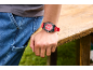 Mão de homem com relógio vermelho, apoiada numa mesa de madeira, com jeans azuis.
