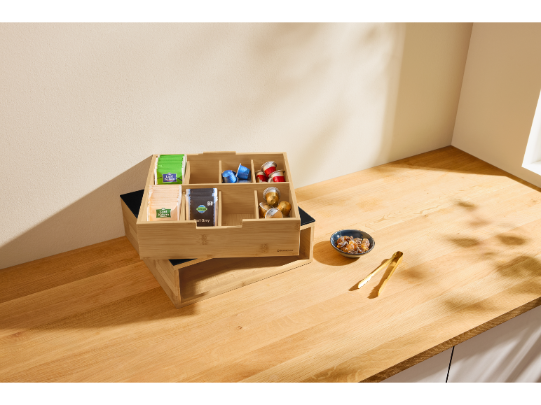 Organizador de madeira para chá e café com saquetas de chá e cápsulas numa mesa de madeira.
