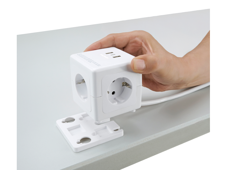 Mão segurando uma extensão cúbica branca com tomadas e portas USB, fixada a um suporte de mesa.