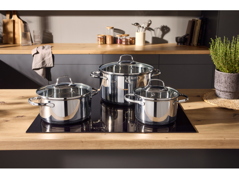 Três panelas de aço inoxidável com tampas de vidro numa placa de indução numa cozinha moderna.