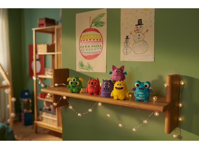 Seis brinquedos de monstros coloridos numa prateleira de madeira num quarto de criança com desenhos de Natal.