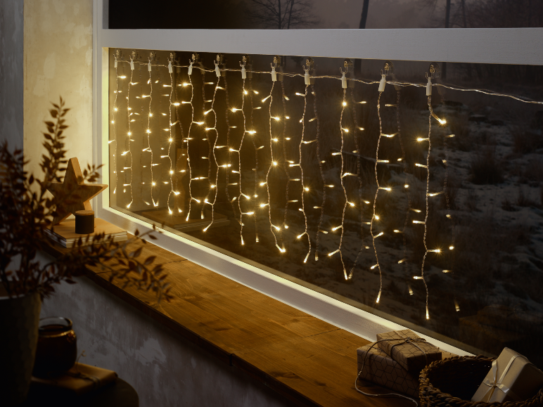Luzes de corda tipo cascata numa janela com decoração de madeira e presentes, criando um ambiente acolhedor.