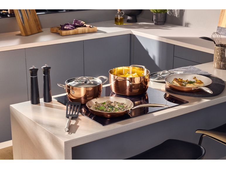 Panelas e frigideiras de cobre numa placa de indução, com comida e acessórios de cozinha.
