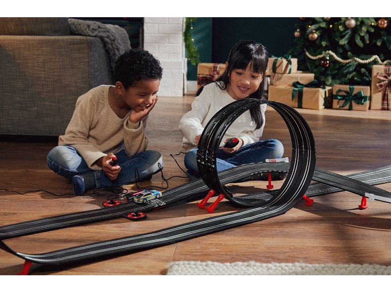 Duas crianças a brincar com uma pista de carros de brinquedo com loops, com presentes de Natal ao fundo.