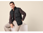 Homem sorridente com colete acolchoado preto e camisola castanha, sentado num cubo branco.