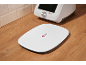 Balança de cozinha digital branca com logótipo numa bancada de madeira, com um robot de cozinha ao fundo.