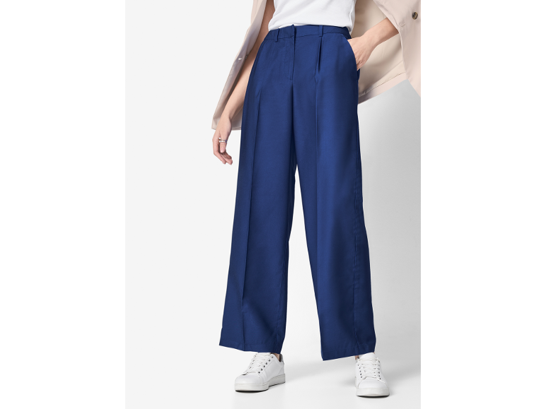 Mulher com calças azuis de perna larga, top branco, blazer bege e ténis brancos.