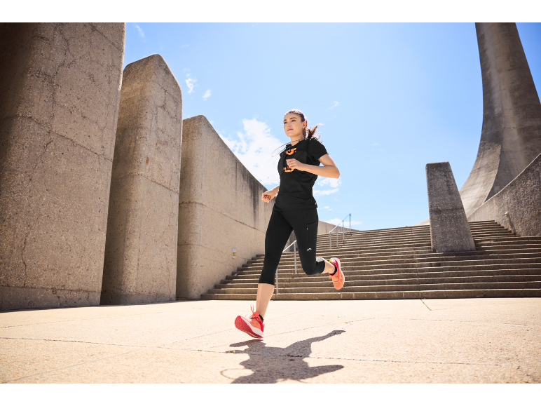Mulher de roupa desportiva preta e sapatilhas de corrida laranja a fazer jogging ao ar livre perto de estruturas de betão.