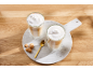 Dois lattes macchiato com espuma de leite, biscoitos e uma colher dourada numa bandeja de mármore.