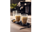 Dois copos de café com espuma e biscoitos numa bandeja, com máquina de café ao fundo.