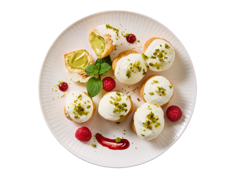 Profiteroles com creme de pistácio, natas, framboesas e hortelã num prato branco.