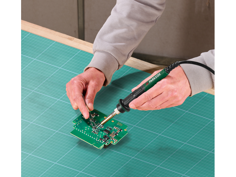 Homem a soldar uma placa de circuito impresso com um ferro de soldar e pinças numa base de corte.