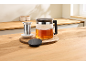 Bule de vidro com chá e um infusor de chá de metal numa bancada de cozinha de madeira.