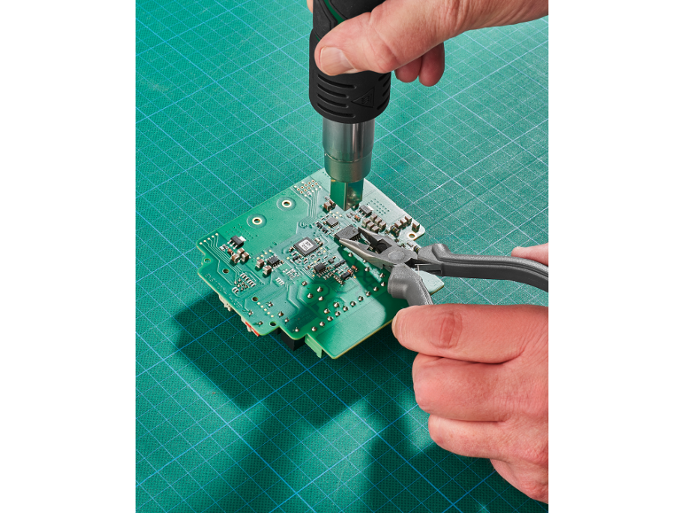 Mãos a reparar uma placa de circuito com uma pistola de ar quente e alicates numa base de corte.