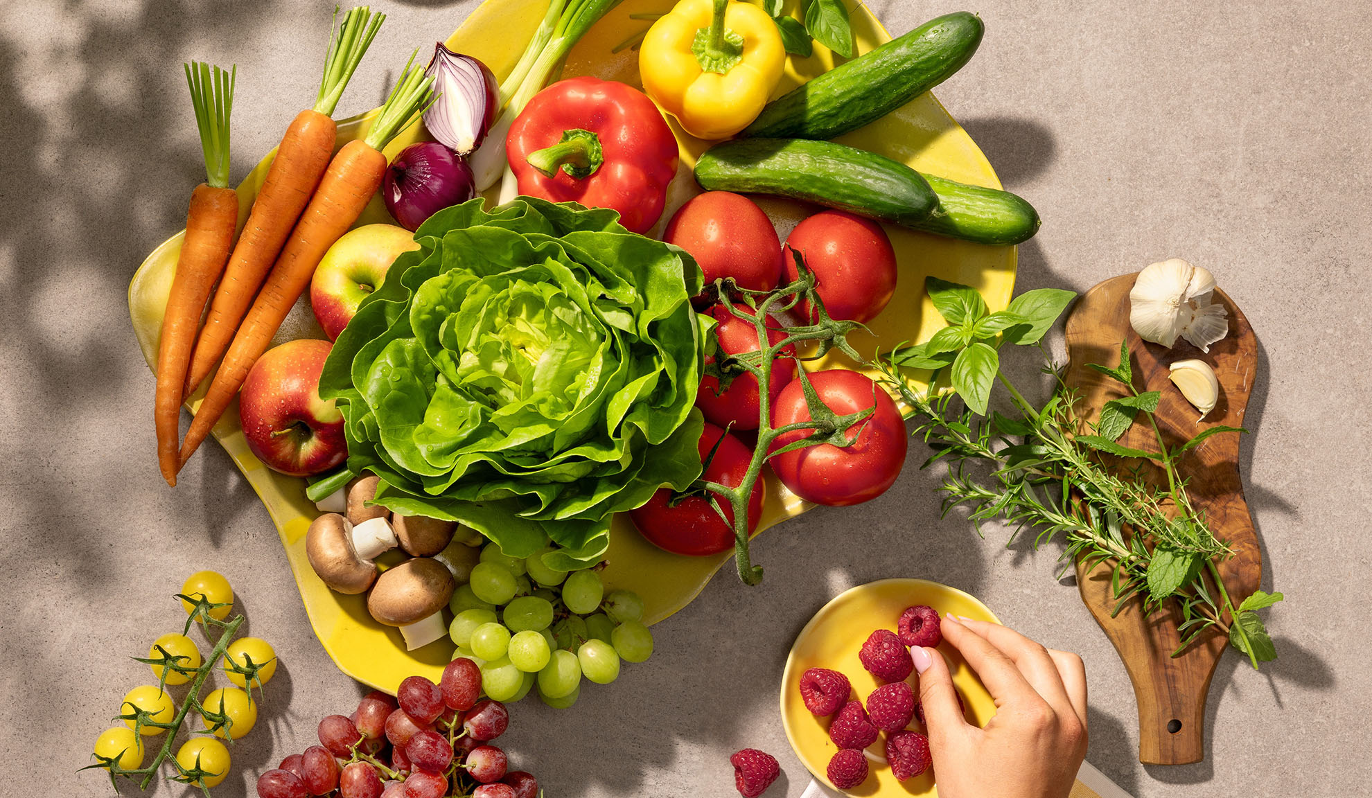 Vegetais e frutas frescas num prato amarelo, incluindo cenouras, alface, pimentões, tomates e uvas.