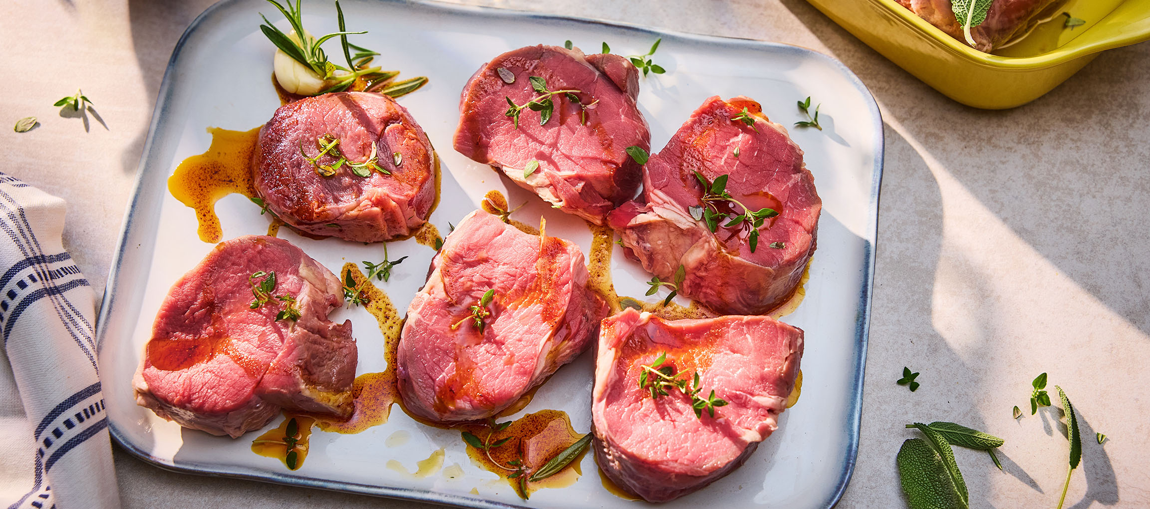 Bifes de carne fresca com ervas e azeite numa travessa branca.
