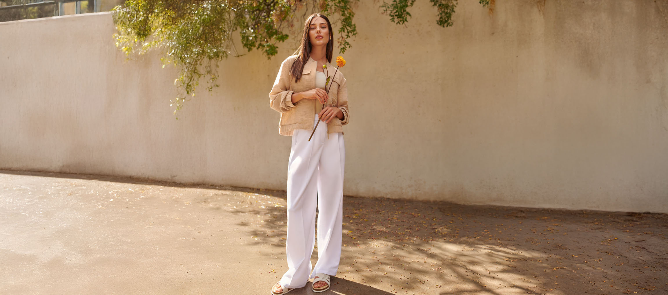 Mulher de casaco bege, calças brancas e sandálias, segurando uma flor amarela.