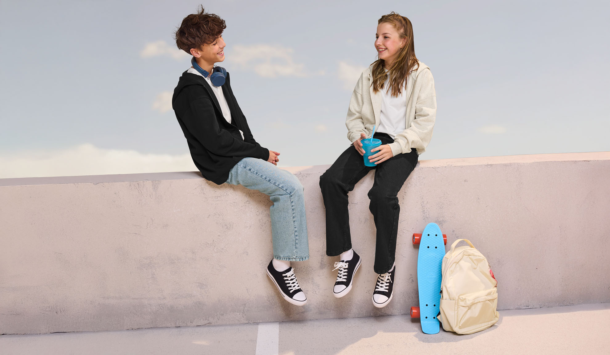 Dois adolescentes com roupa casual, sentados num muro, com um skate e uma mochila por perto.