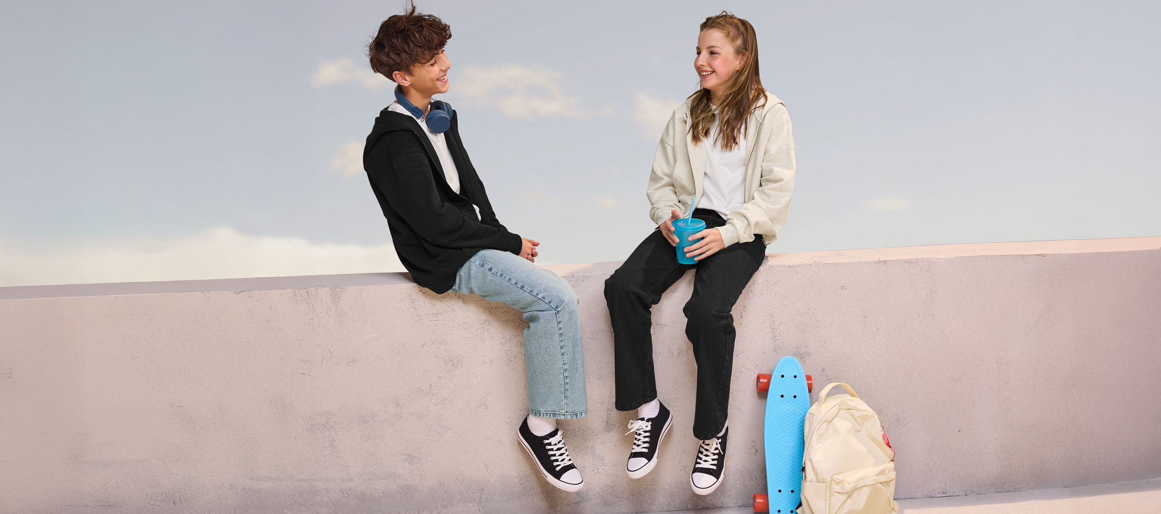 Dois adolescentes de hoodies e jeans sentados num muro, com um skate e mochila por perto.