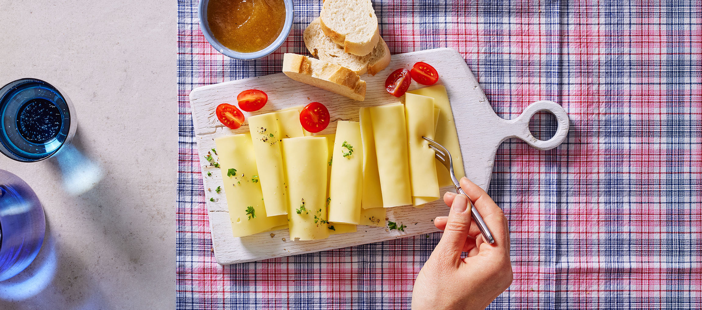 Fatias de queijo com tomate cereja, pão e geleia sobre uma toalha xadrez.
