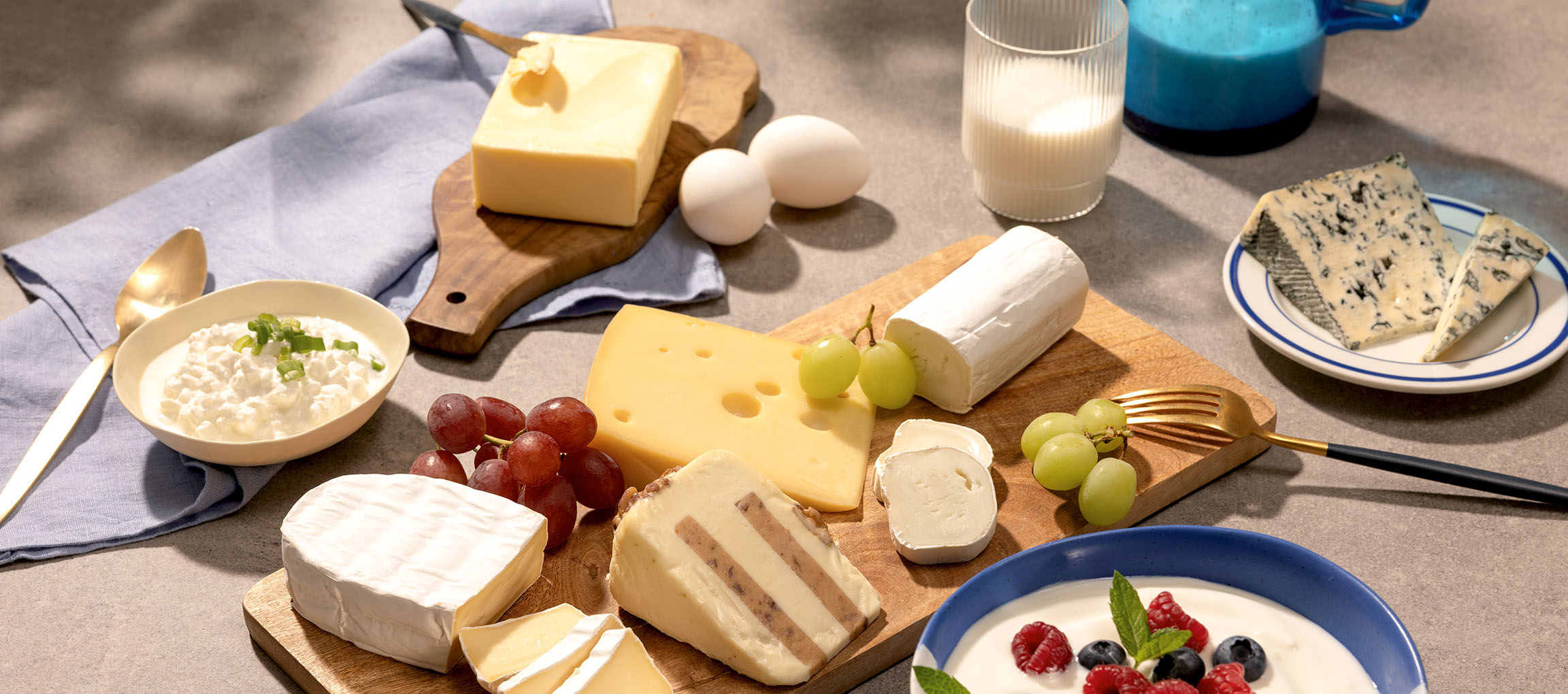 Variedade de queijos, ovos, uvas, leite e iogurte com frutos silvestres numa mesa.