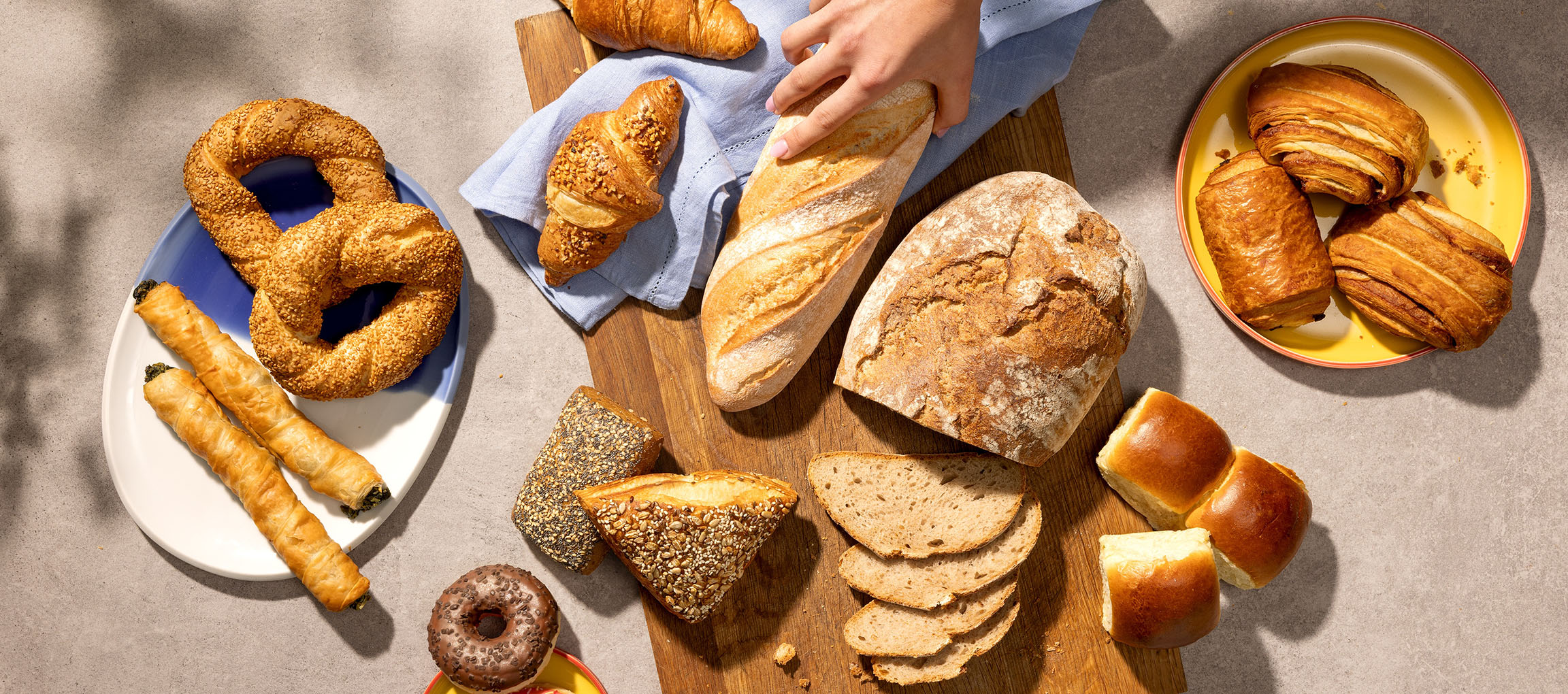 Variedade de pastelaria e pão, incluindo pretzels, croissants e baguetes, numa mesa.