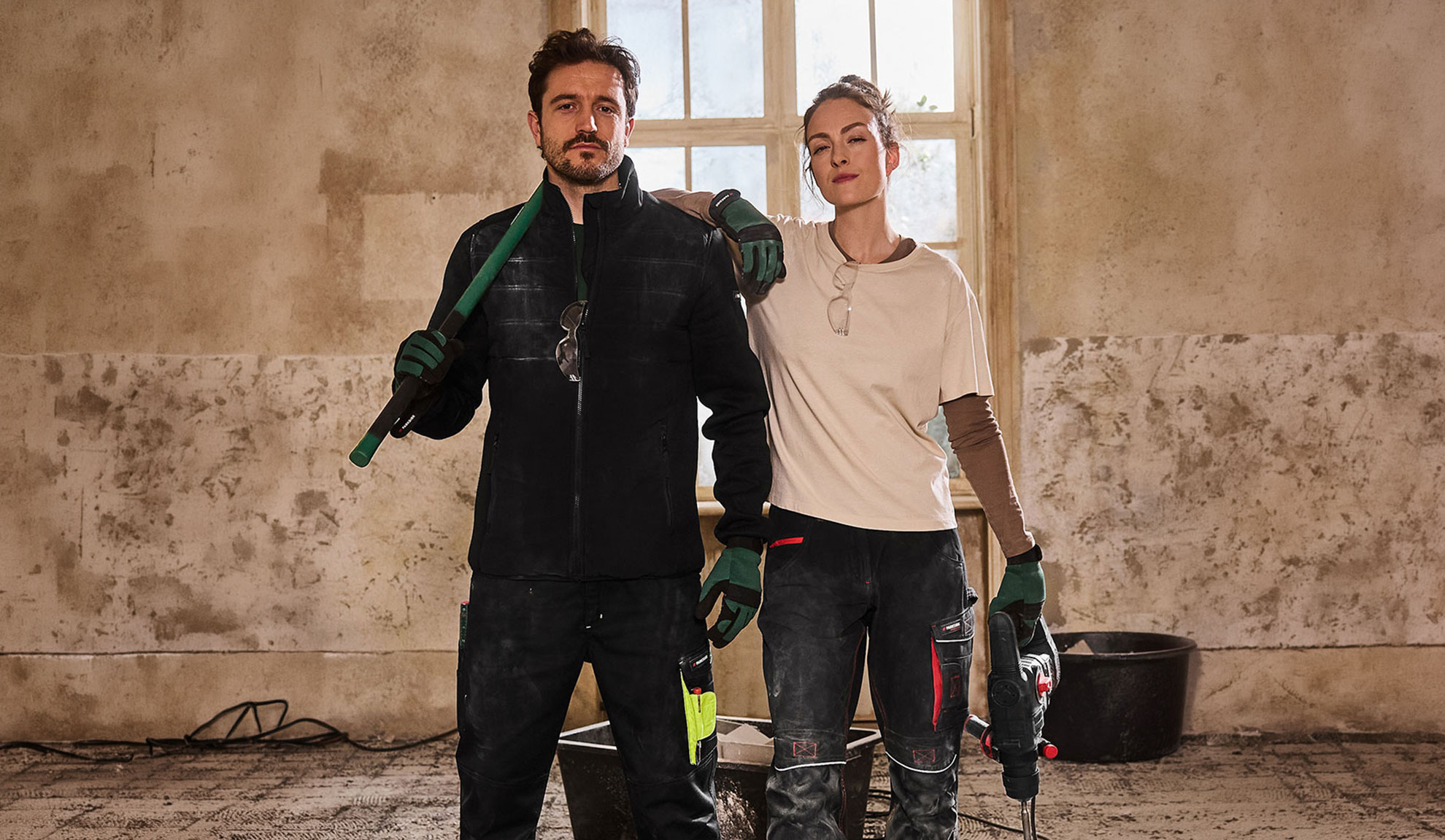 Dois trabalhadores da construção civil em roupa de trabalho, homem com pá e mulher com berbequim.