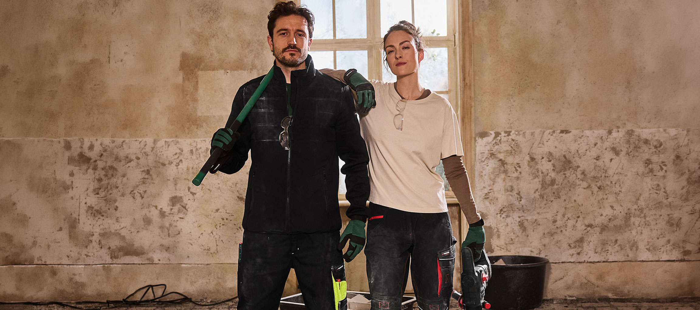 Homem e mulher em roupa de trabalho, com ferramentas, numa sala em renovação.
