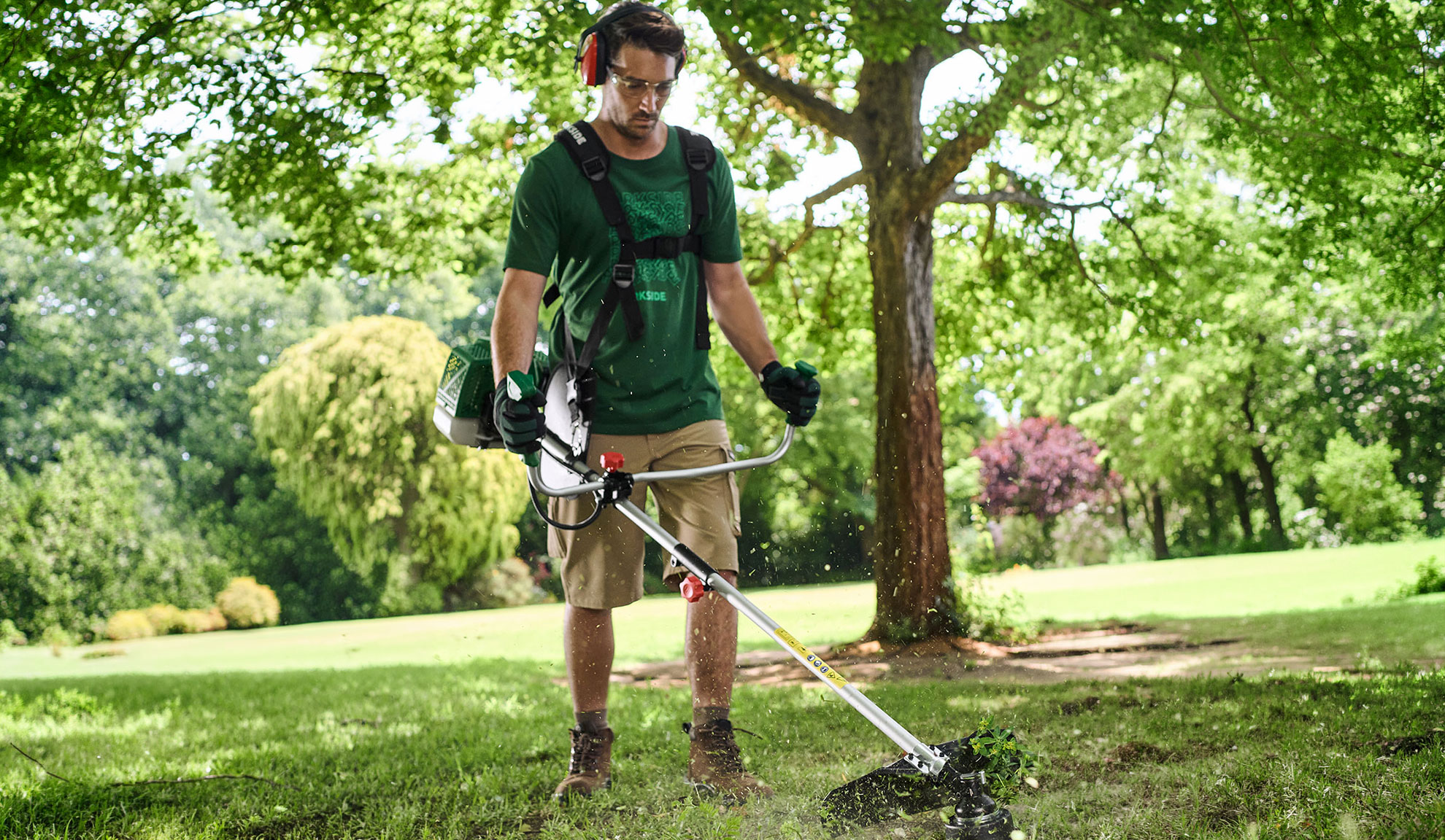 Homem a cortar relva num jardim com uma roçadora, usando óculos de segurança e protetores auriculares.