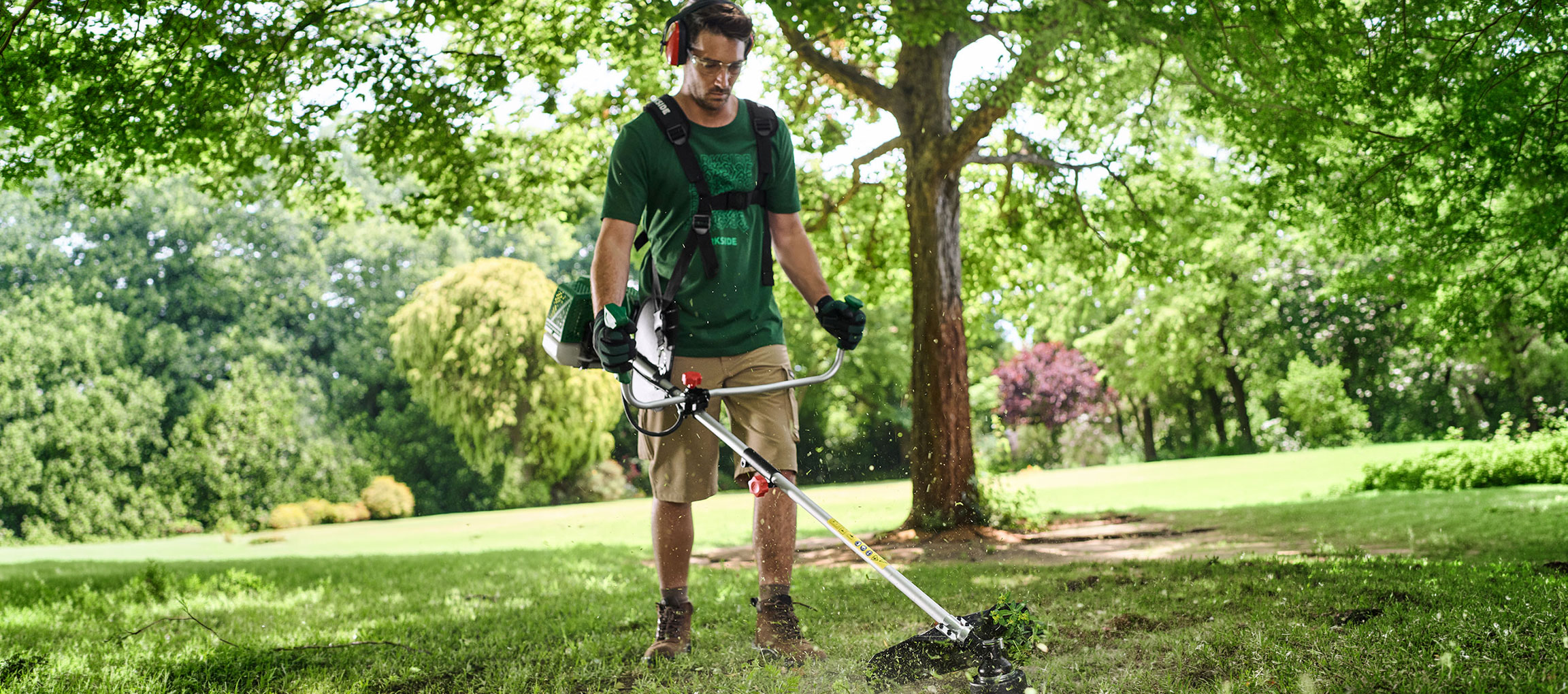 Homem com roçadora e equipamento de proteção a cortar relva num jardim.