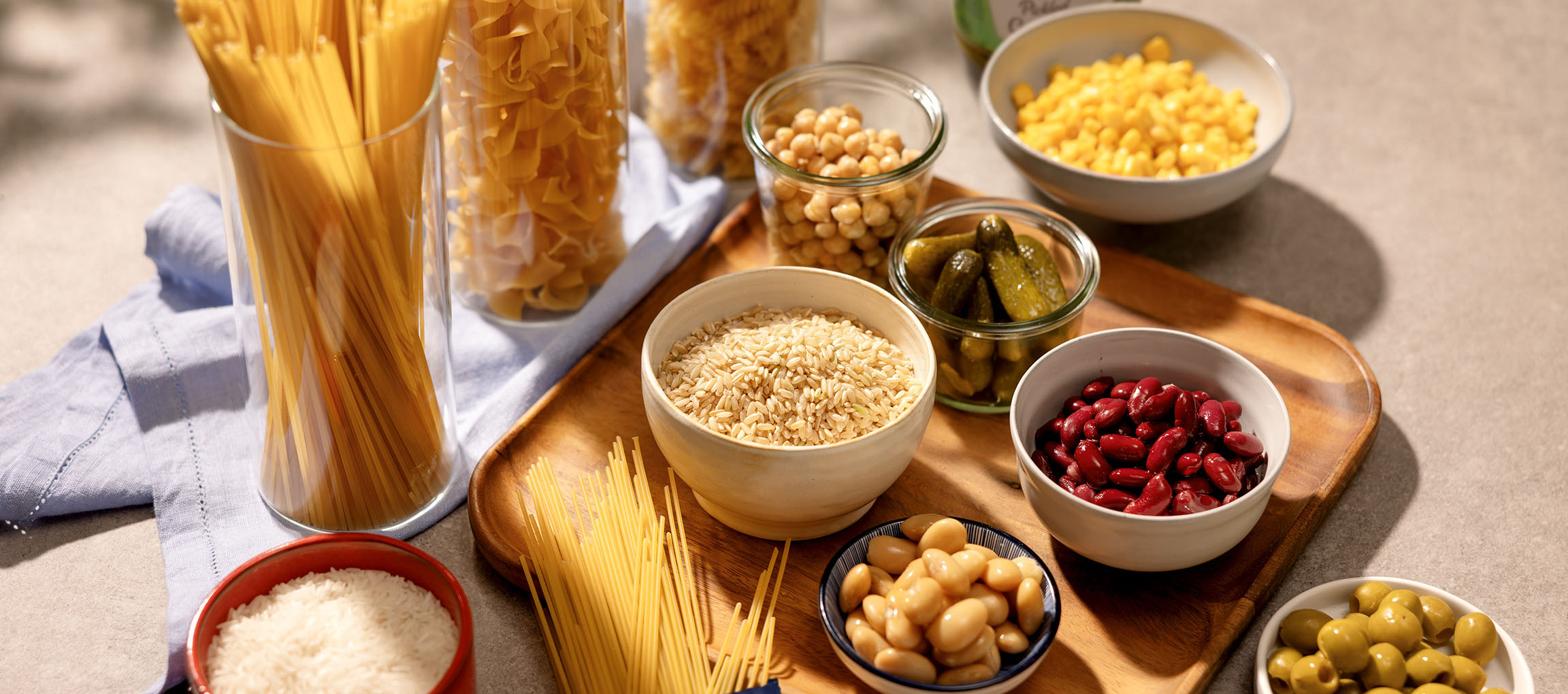 Vários produtos básicos de despensa, incluindo massa, arroz, feijão, grão-de-bico, milho, picles e azeitonas.