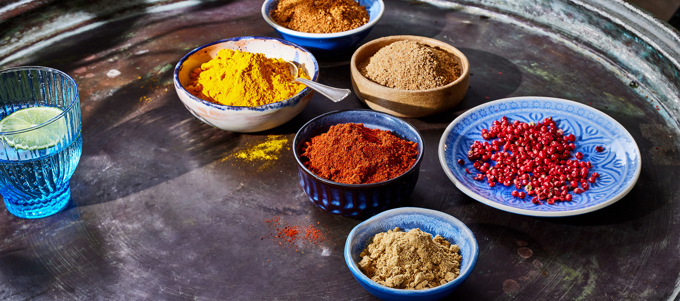 Várias especiarias, como açafrão, pimentão e pimenta rosa, em tigelas sobre uma superfície escura.