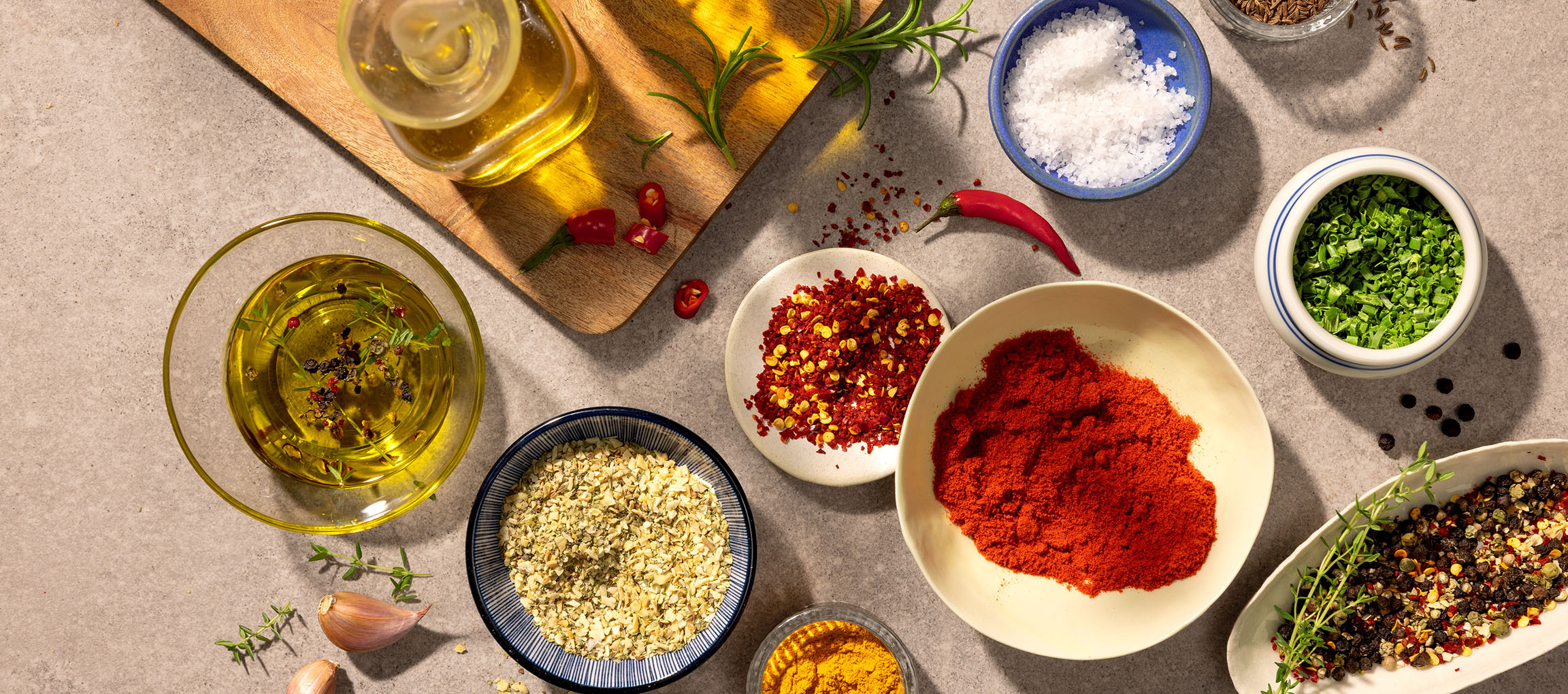 Variedade de especiarias e azeites, incluindo pimentão, chili, sal, pimenta e azeite, sobre fundo cinzento.