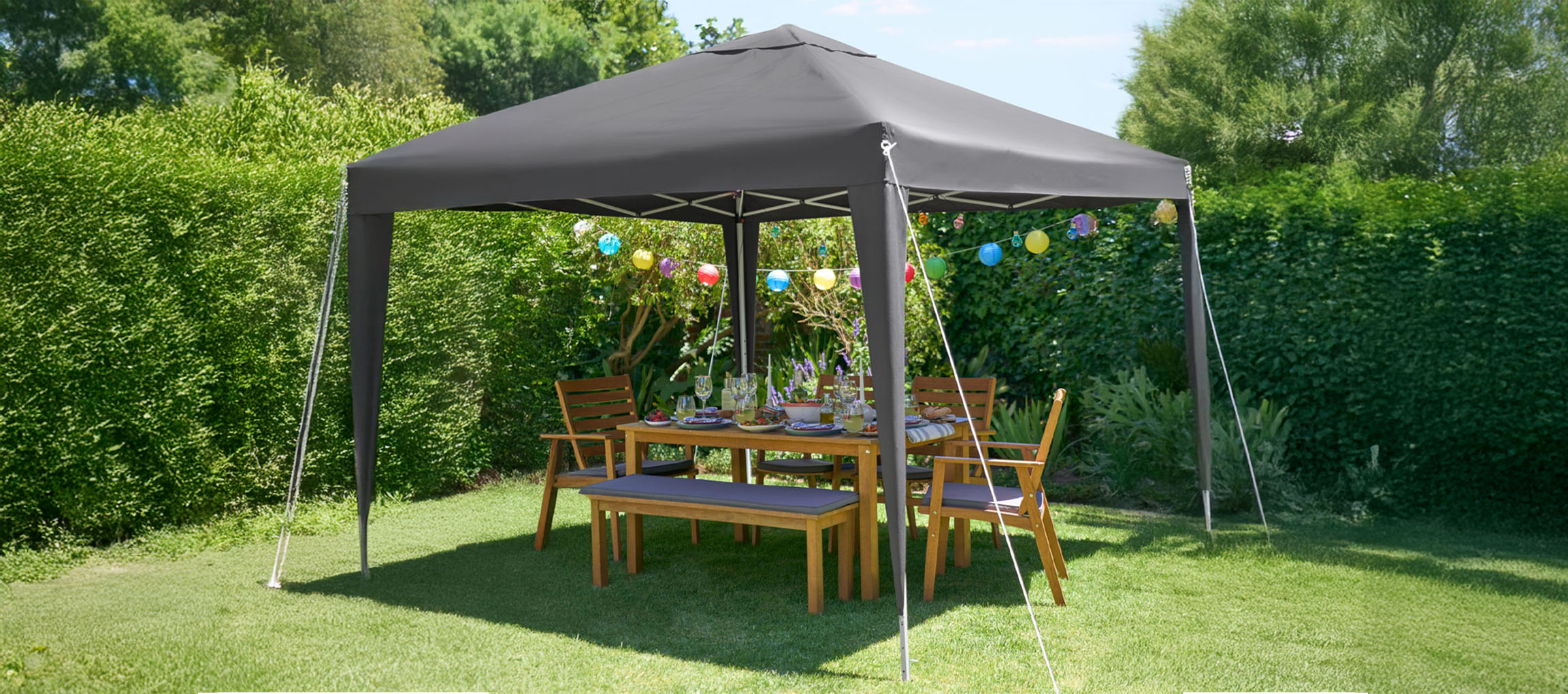 Um gazebo retrátil cinza-escuro protege um conjunto de jantar de madeira para exterior num jardim verdejante.