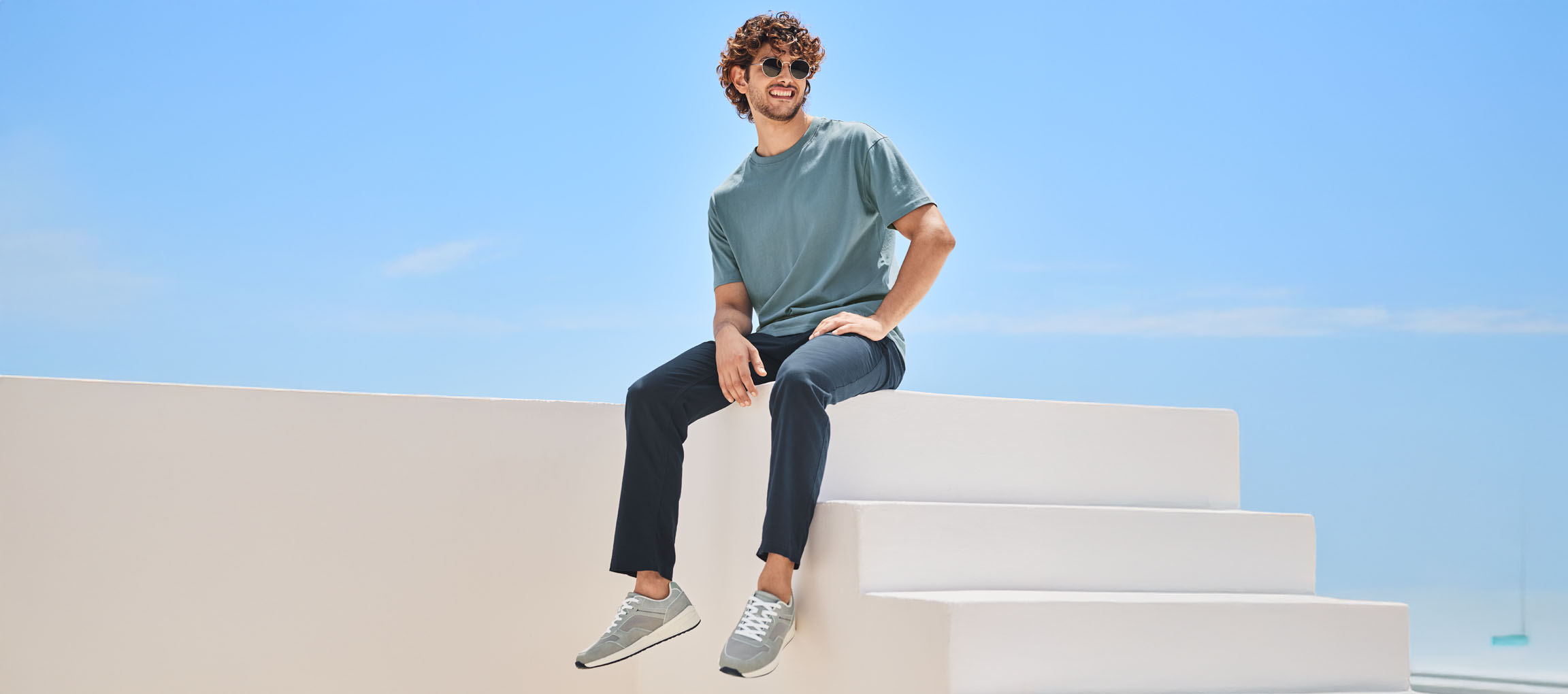 Homem relaxado vestindo camiseta, calça e tênis New Balance, sentado em uma escada branca.