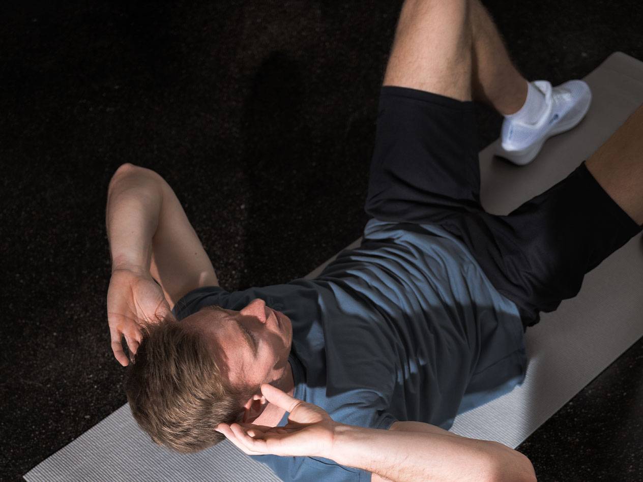 Homem de camiseta azul e shorts pretos fazendo abdominais em um tapete de yoga.