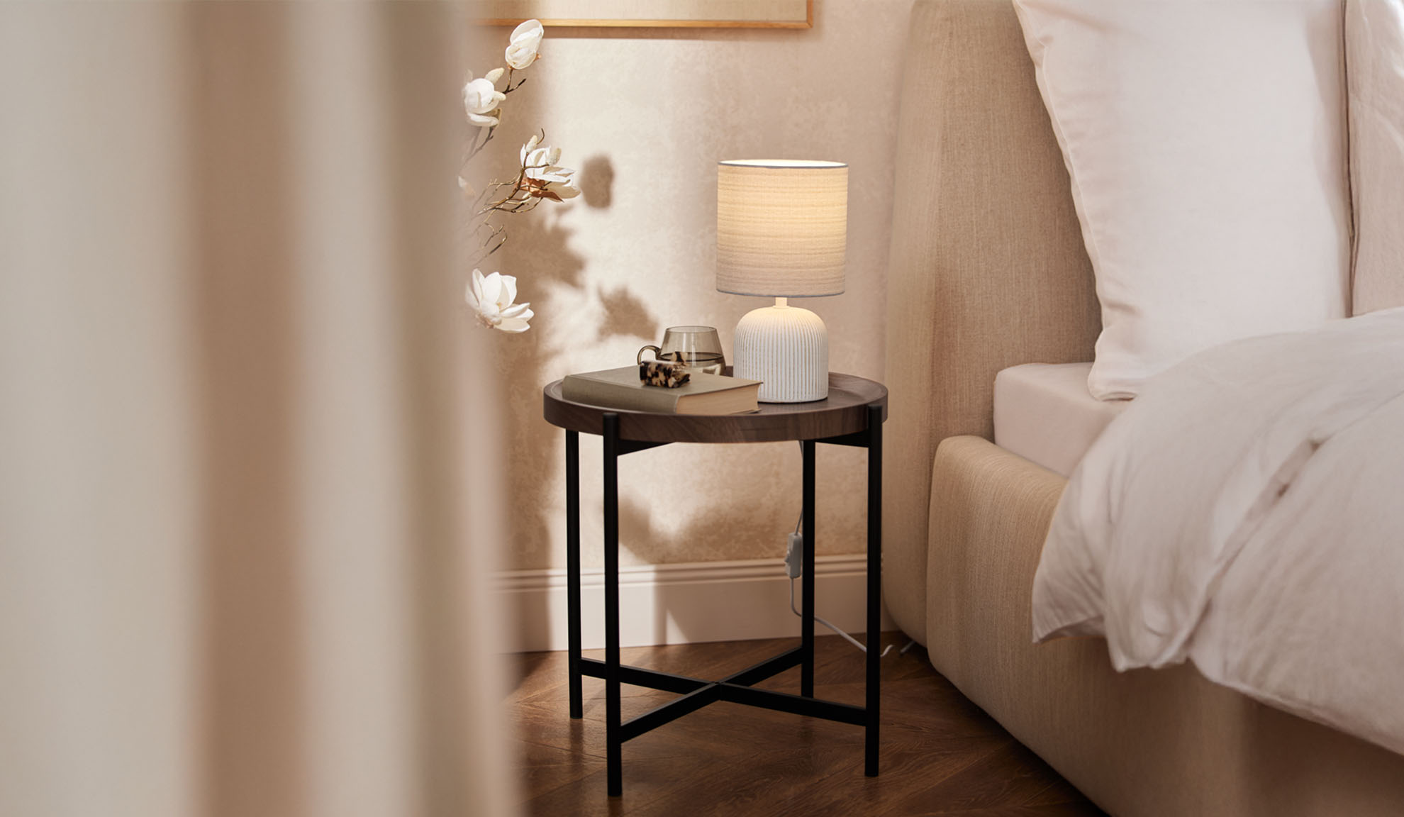 Mesa de cabeceira redonda de madeira com candeeiro de mesa e acessórios ao lado de uma cama.