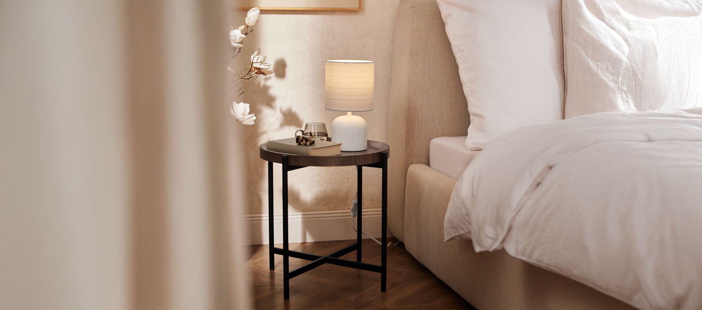 Mesa de cabeceira redonda de madeira com candeeiro de mesa e cama bege num quarto acolhedor.