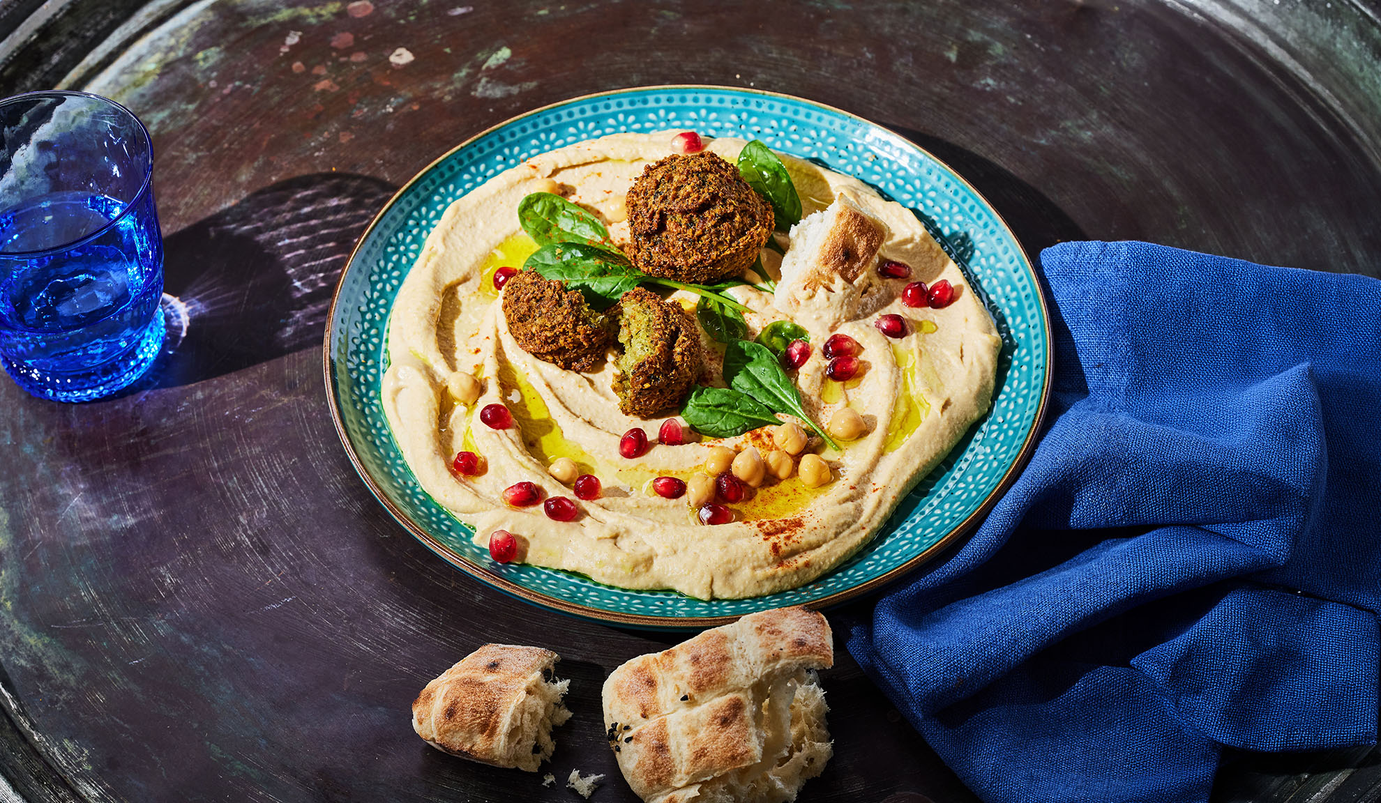 Hummus com falafel, sementes de romã e pão pita numa mesa escura.