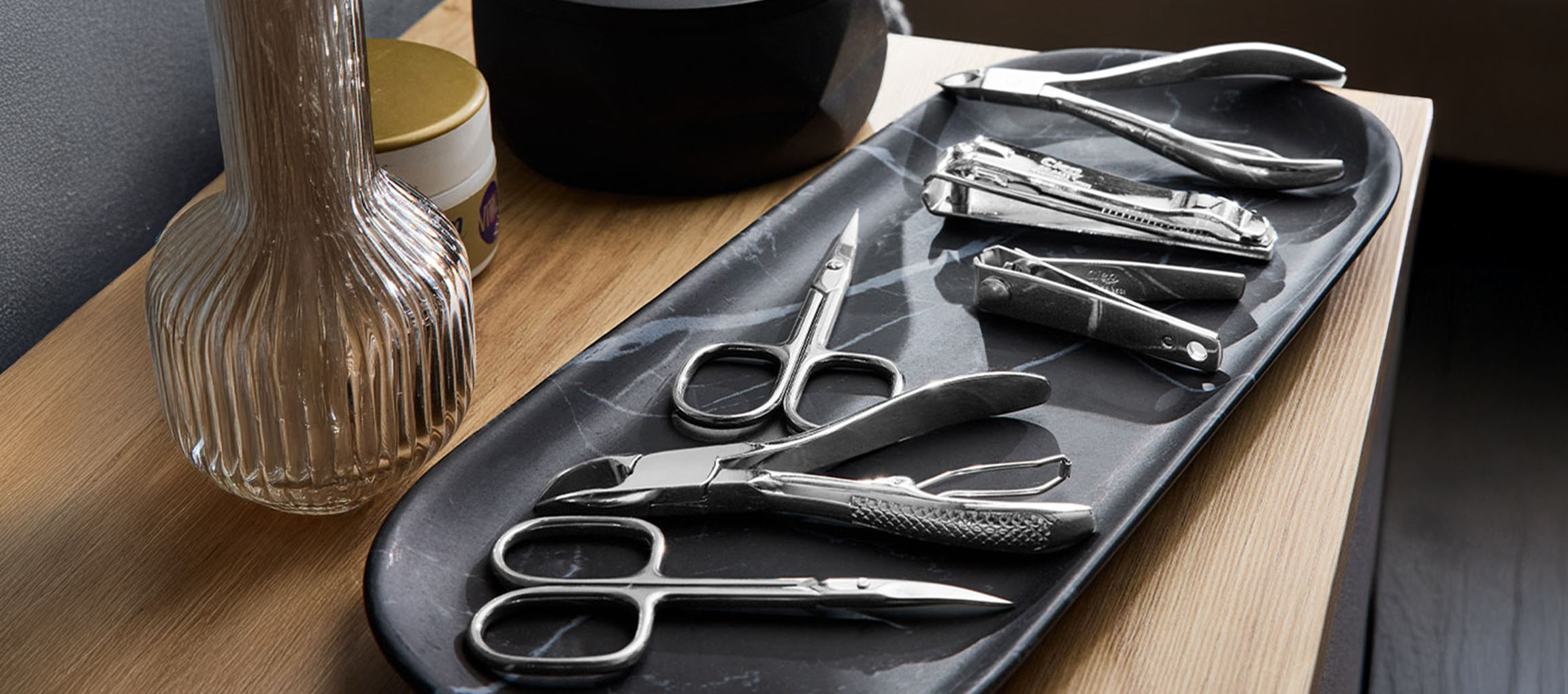 Ferramentas de manicure e pedicure numa bandeja de mármore, com um vaso e frasco ao fundo.