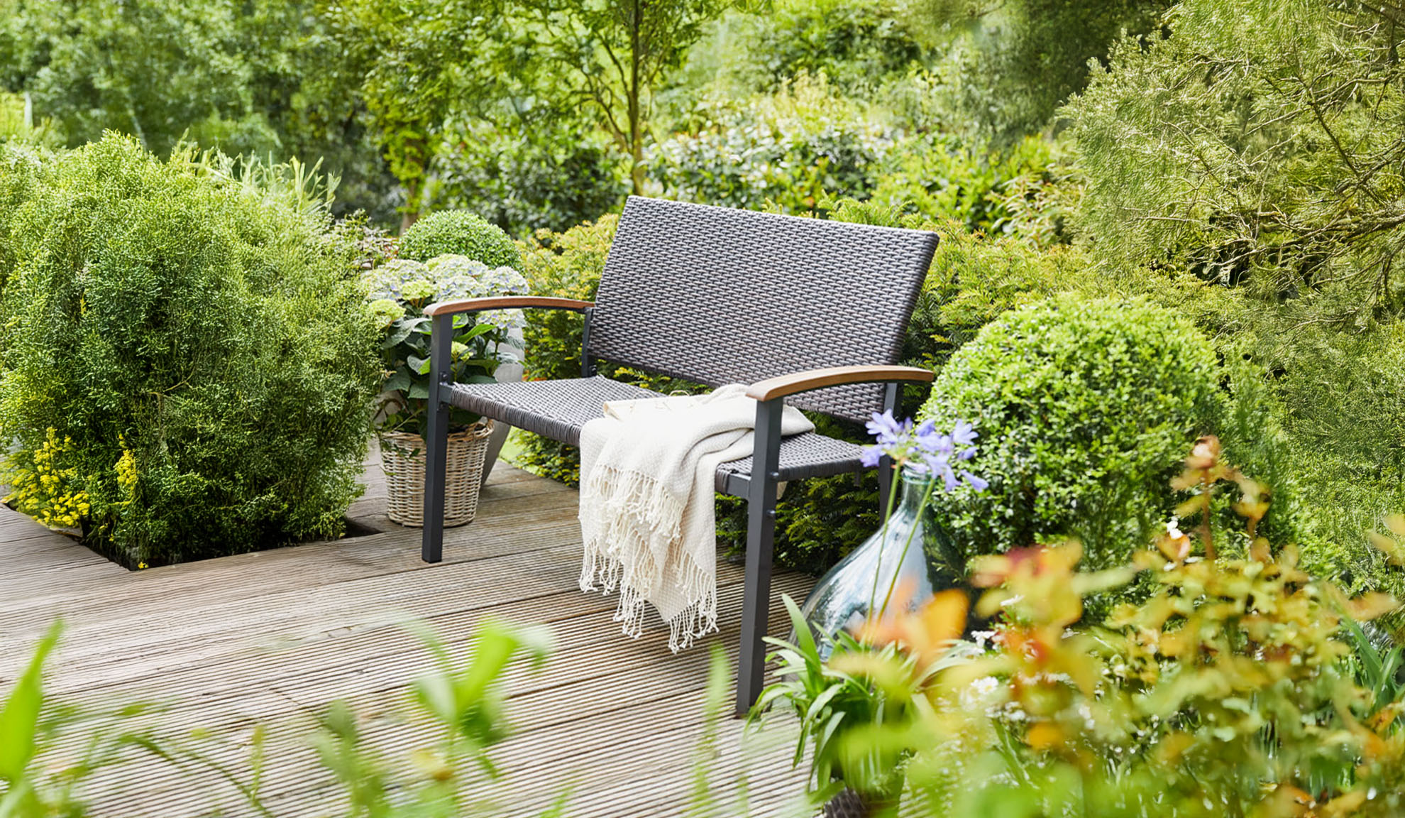 Banco de jardim de vime em um deck de madeira, cercado por vegetação exuberante.
