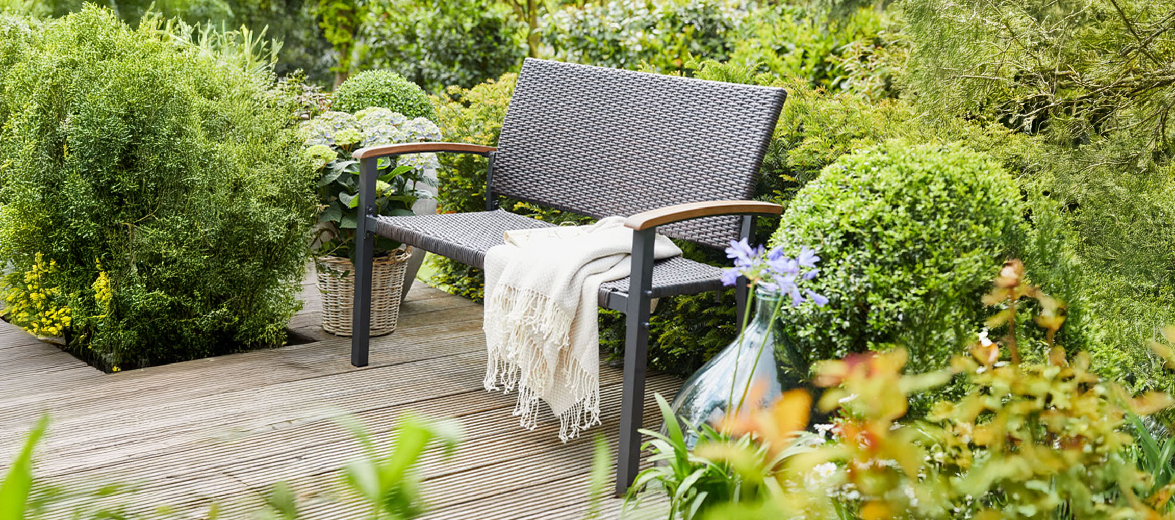 Um banco de jardim cinzento com um cobertor num deck de madeira rodeado por plantas verdes.