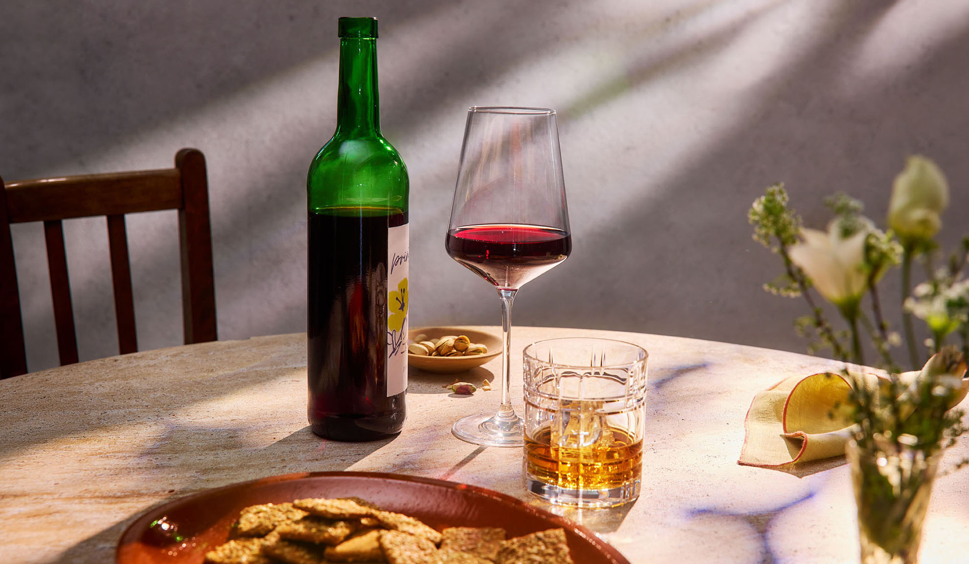 Vinho tinto, uísque e petiscos numa mesa à luz do sol.