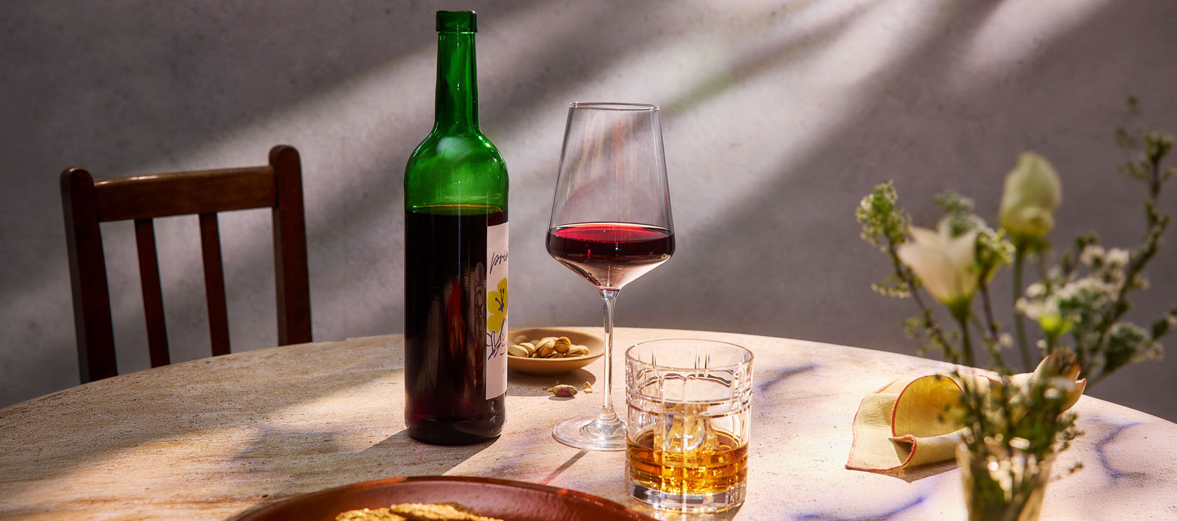 Garrafa de vinho tinto, copo de vinho e uísque numa mesa, com cadeira e flores ao fundo.