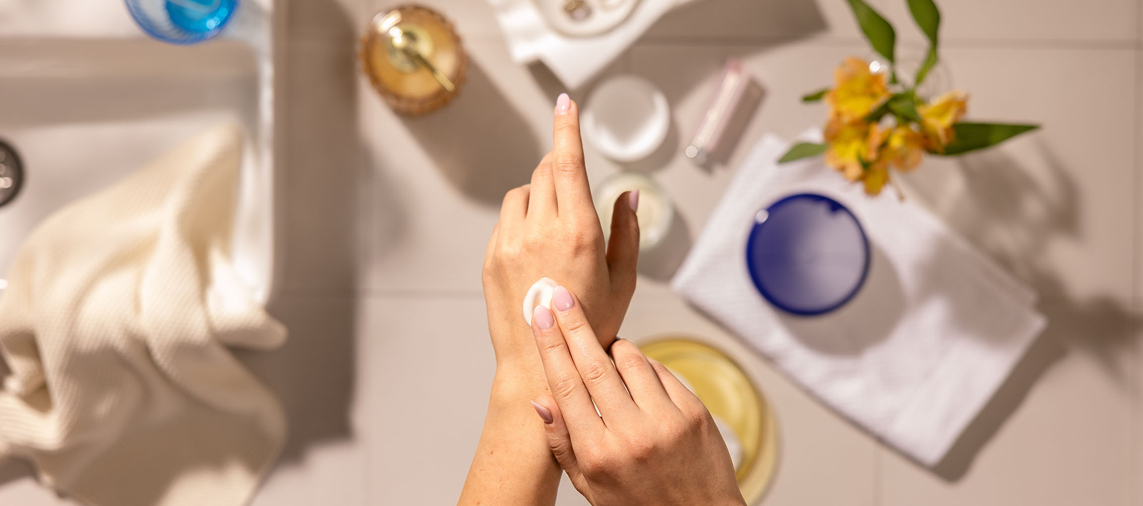 Mãos femininas aplicando creme para as mãos, rodeadas por cosméticos e toalhas numa casa de banho.